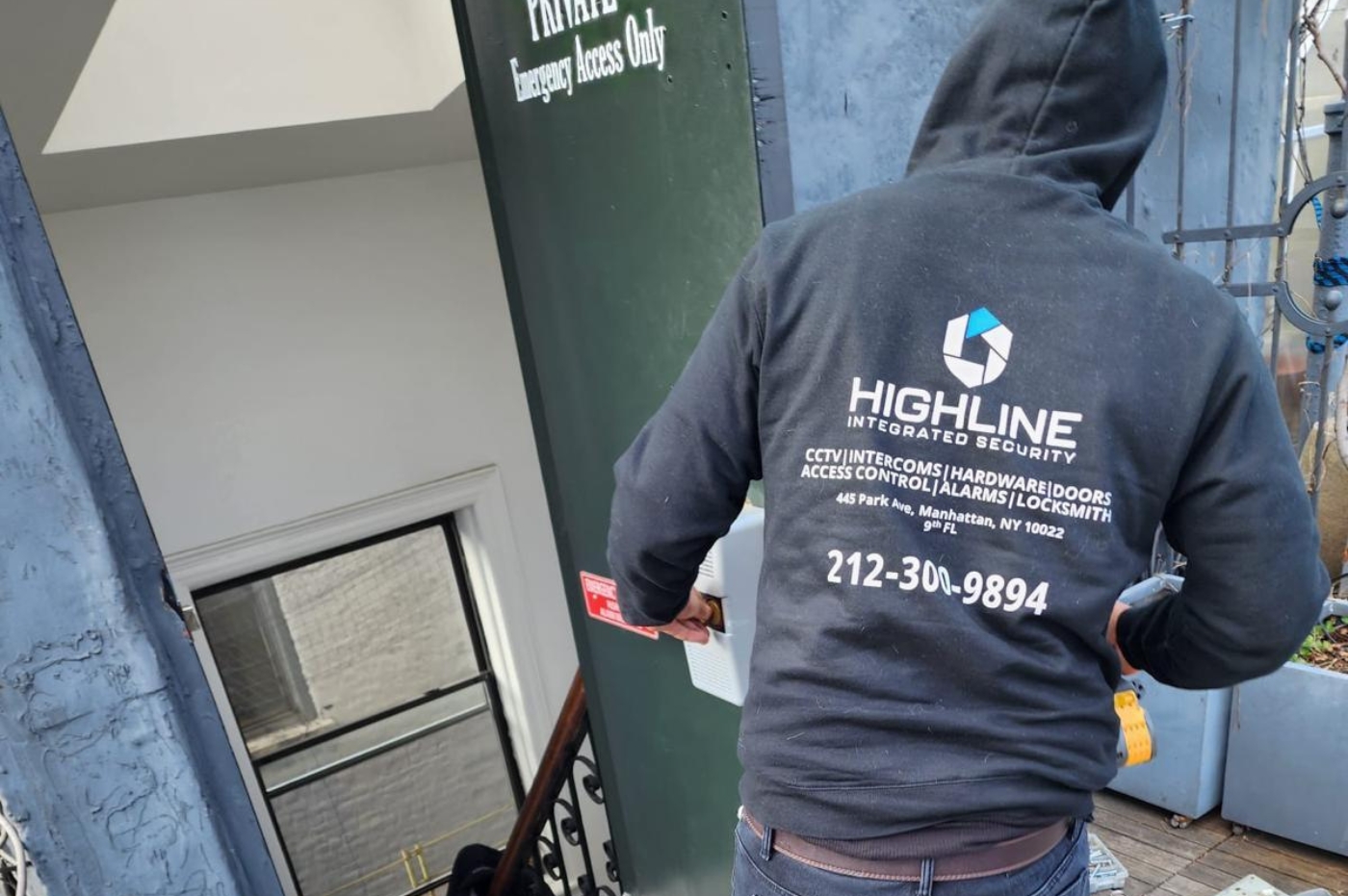 A person in a "HIGHLINE INTEGRATED SECURITY" hoodie works near a door labeled "PRIVATE Emergency Access Only." The hoodie also lists services like CCTV and locksmithing with a phone number.