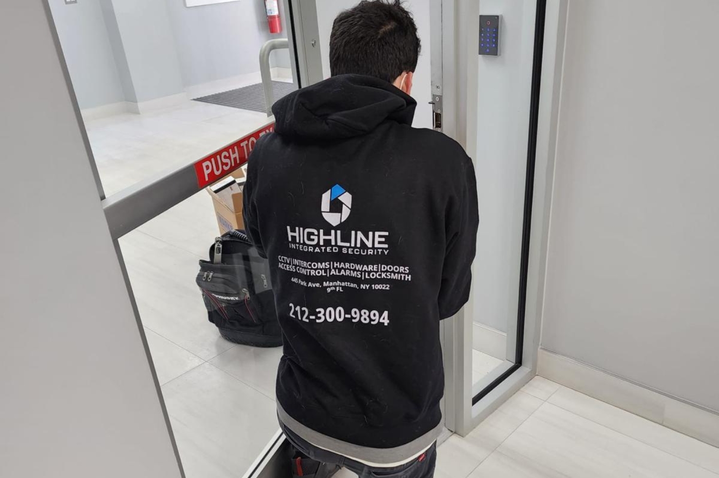 A person in a black hoodie labeled "HIGHLINE INTEGRATED SECURITY" works on a door with an access keypad beside it. Nearby, a push bar and a backpack are visible. Text on hoodie: HIGHLINE INTEGRATED SECURITY CCTV | INTERCOMS | HARDWARE | DOORS ACCESS CONTROL | ALARMS | LOCKSMITH 445 Park Ave, Manhattan, NY 10022 9th FL 212-300-9894