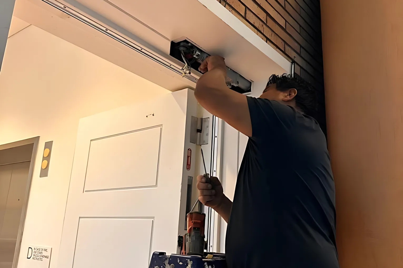 A person is fixing an elevator door mechanism using tools, set in a building corridor with visible brick and wood walls. Elevator buttons and a sign are present.