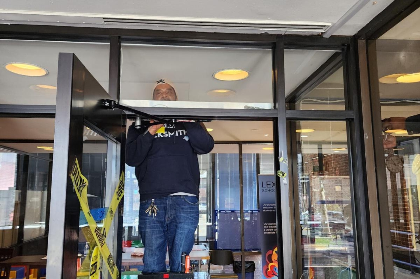 A man standing on a stool adjusts an overhead door closer inside a building, bordered by glass walls. Yellow caution tape crosses the doorway. Keys hang from his jeans.