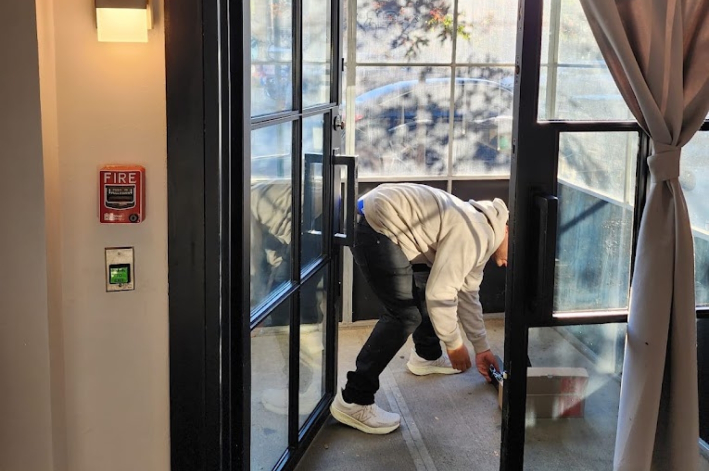 A person in a hooded sweatshirt bends to pick up an object on a sunlit porch. Nearby, a "FIRE" alarm and green button are mounted on the wall.