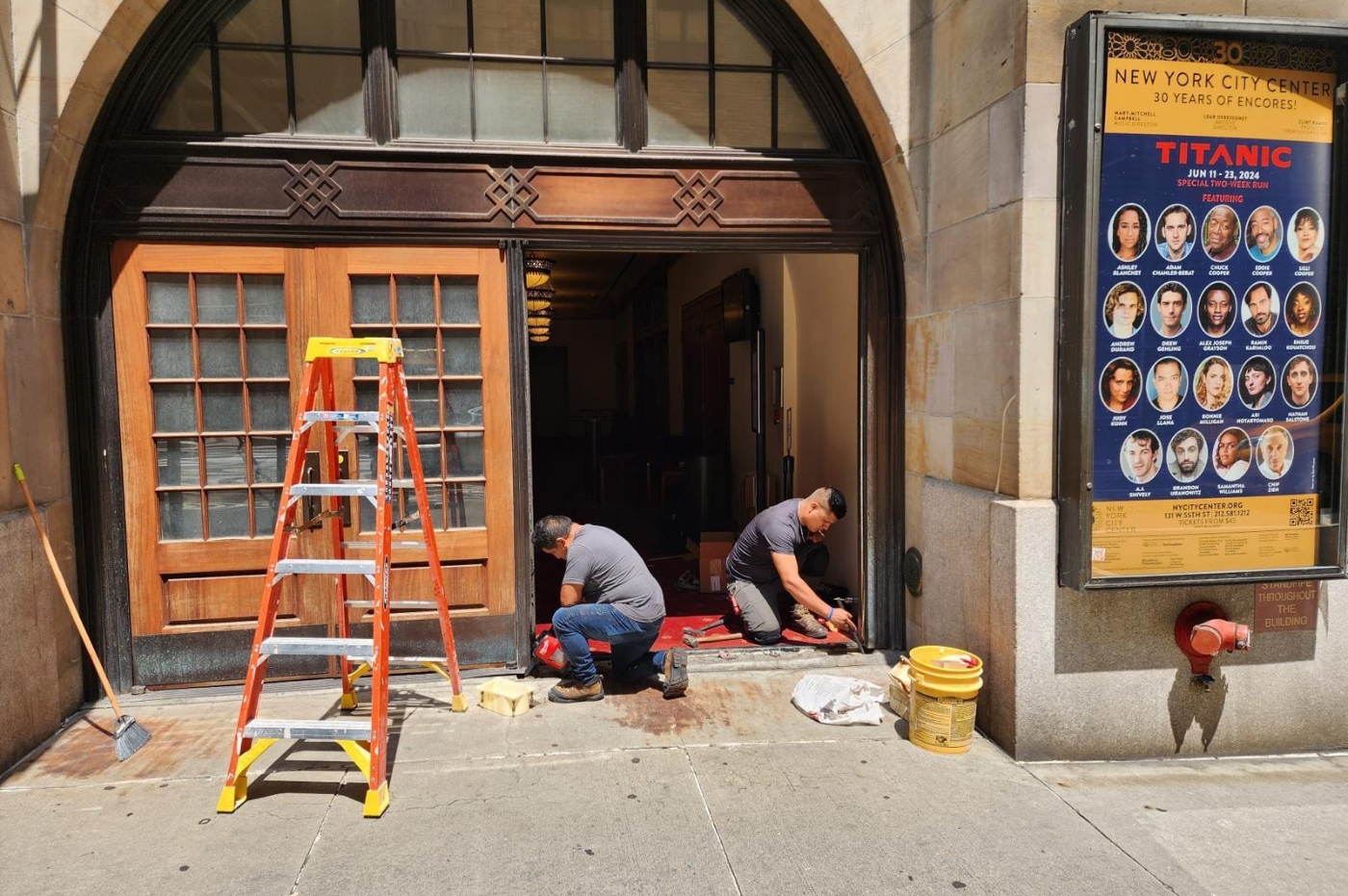 Two workers kneel and work on door repairs at a building entrance under sunlight. An orange ladder stands nearby. A poster advertises "Titanic" at New York City Center, June 11-23, 2024.