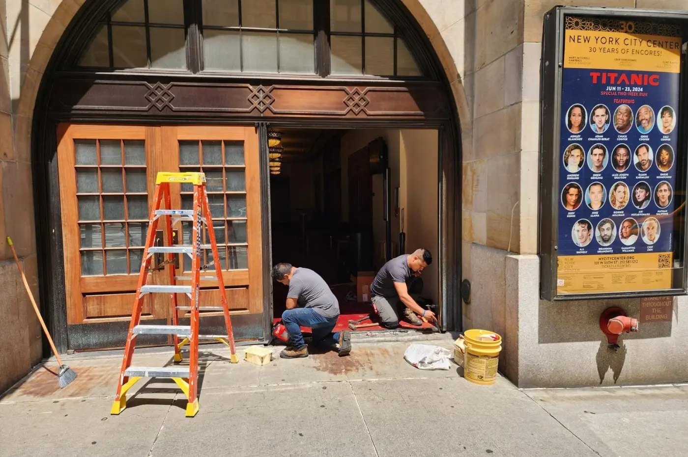 Two workers kneel and work on door repairs at a building entrance under sunlight. An orange ladder stands nearby. A poster advertises "Titanic" at New York City Center, June 11-23, 2024.