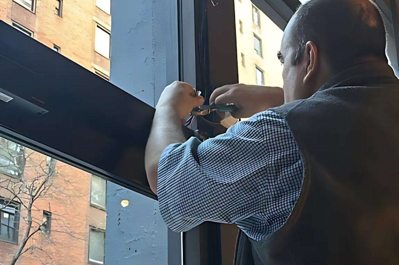 A person uses tools on a window frame inside a building, with an urban street view and brick buildings visible outside.