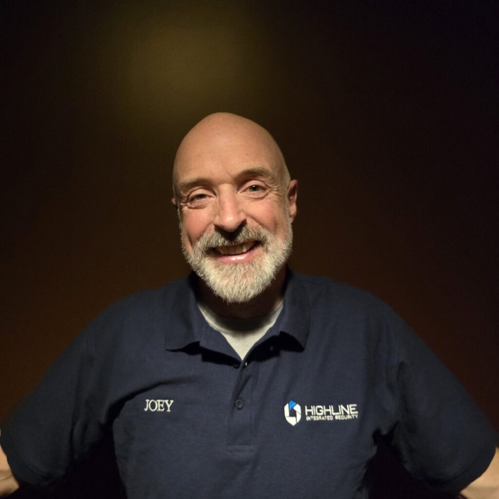 A person smiles with arms outstretched. They wear a dark polo shirt featuring the text “JOEY” and "HIGHLINE INTEGRATED SECURITY." The background is a plain, dark wall.